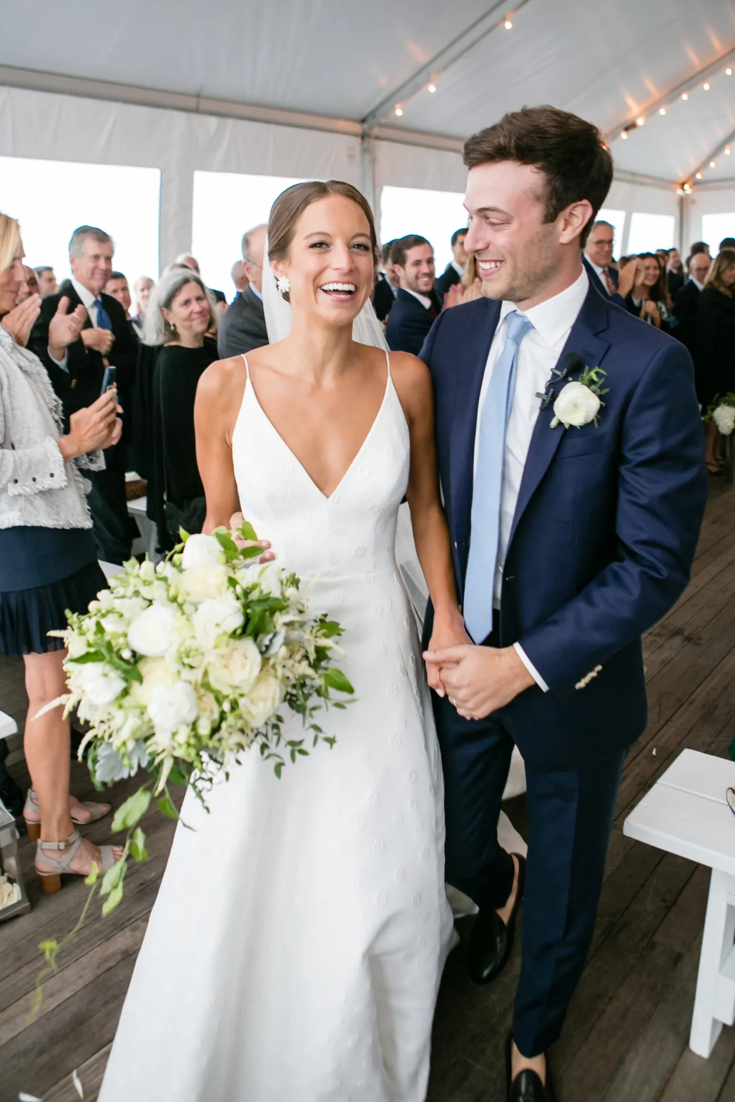 Hannah and Mark walk up the tented aisle laughing, her bouquet of white roses in hand