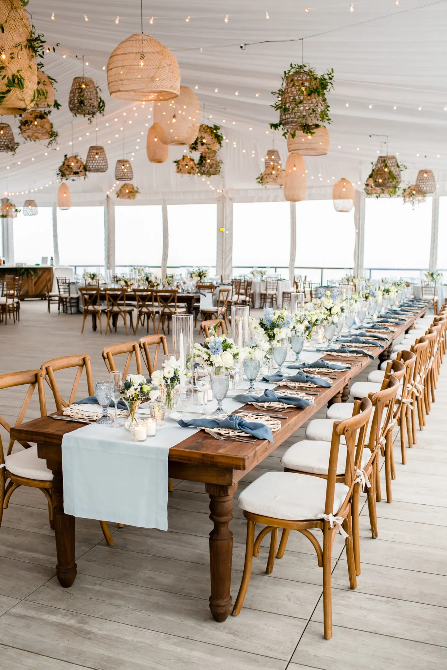 Long wooden farm tables under hanging rattan lanterns and greenery in the reception tent