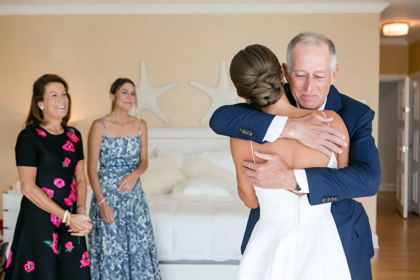 The bride hugs an older man in a navy suit tightly while two women smile in a sunny suite