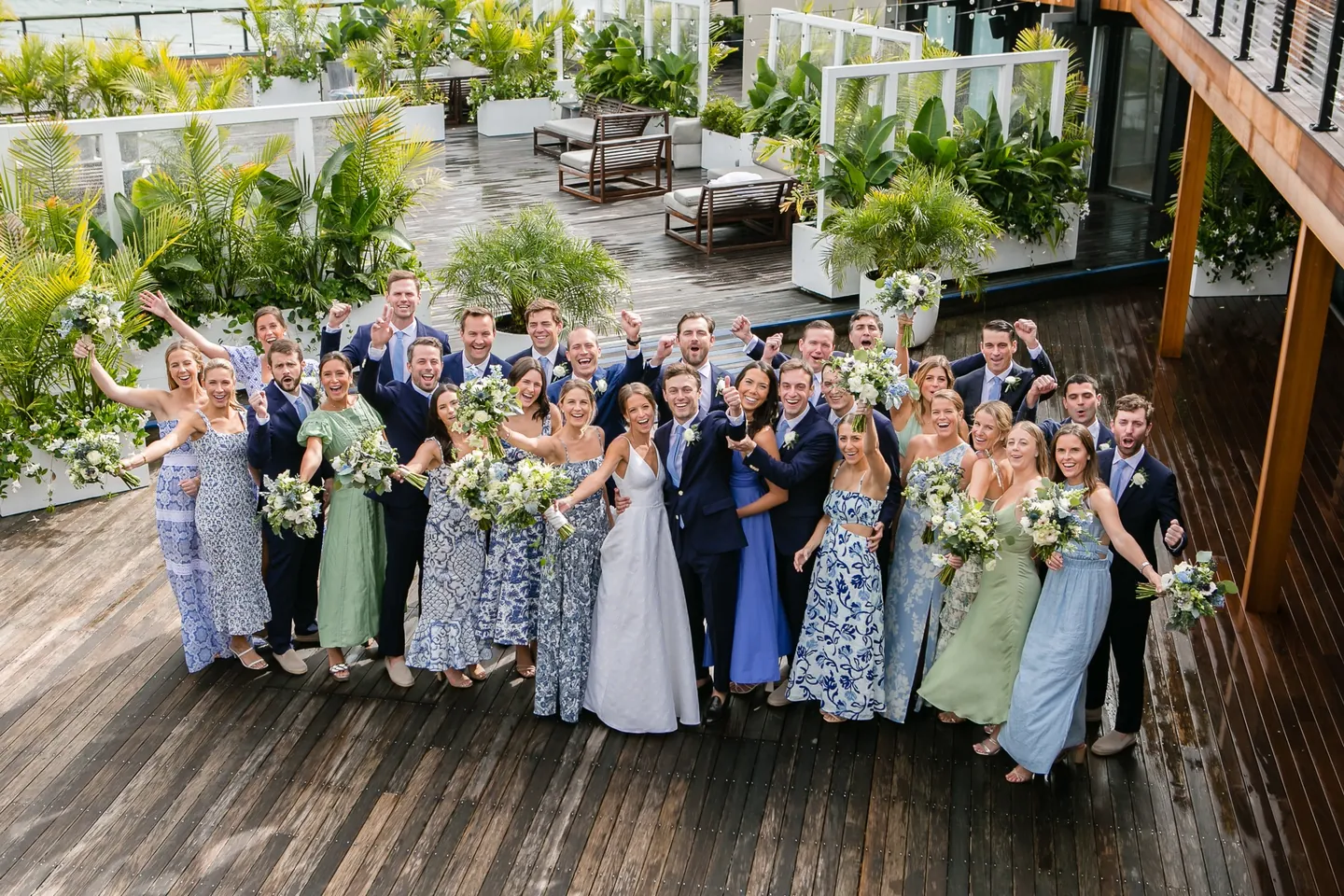 The whole wedding party cheers on a wet wooden deck lined with potted palms and planters