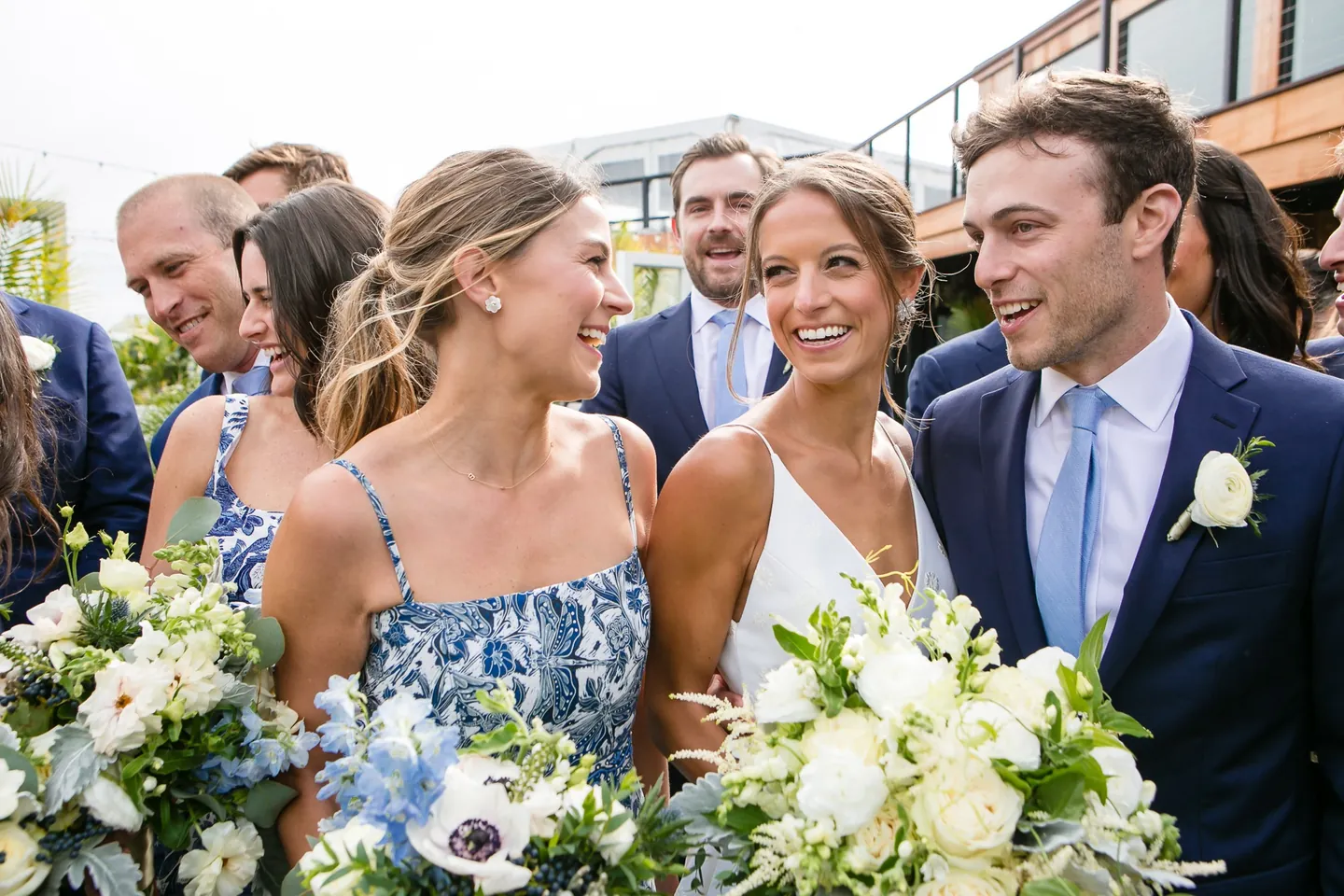 Hannah and Mark laugh close together with bridesmaids and white bouquets around them