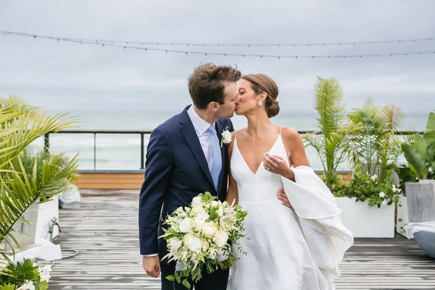 Hannah and Mark kiss on the rooftop deck, the grey ocean and potted palms behind them