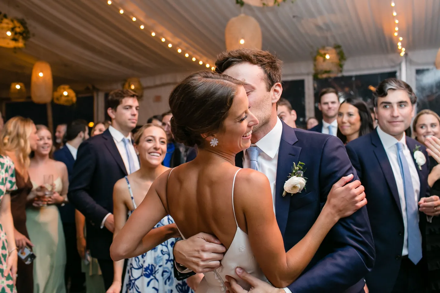 The couple embrace on the dance floor under warm rattan lanterns as guests circle them