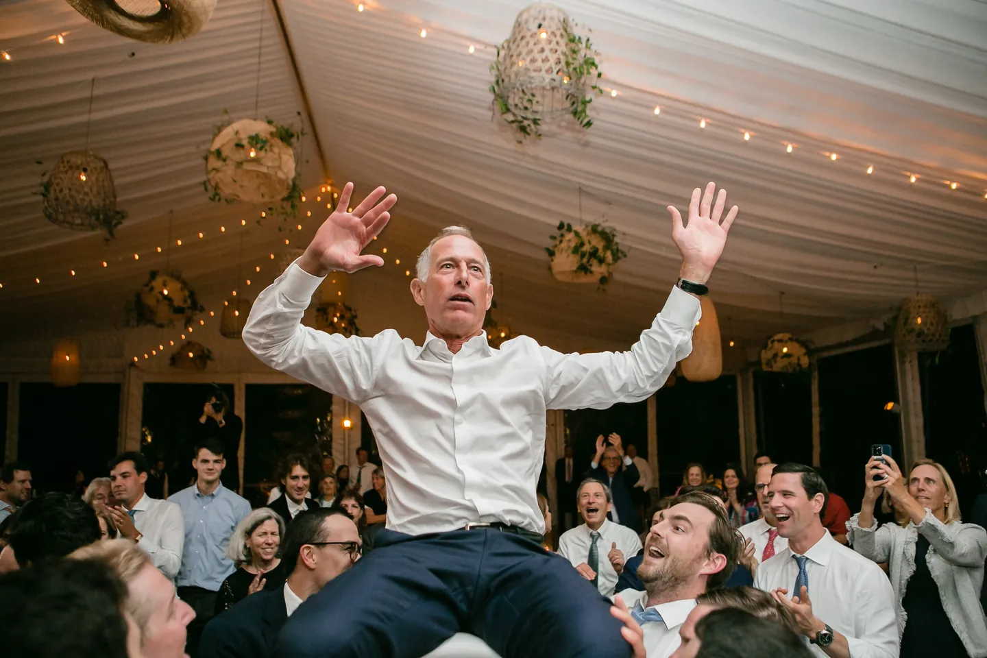 A guest in a white shirt is hoisted in a chair above the crowded tent dance floor
