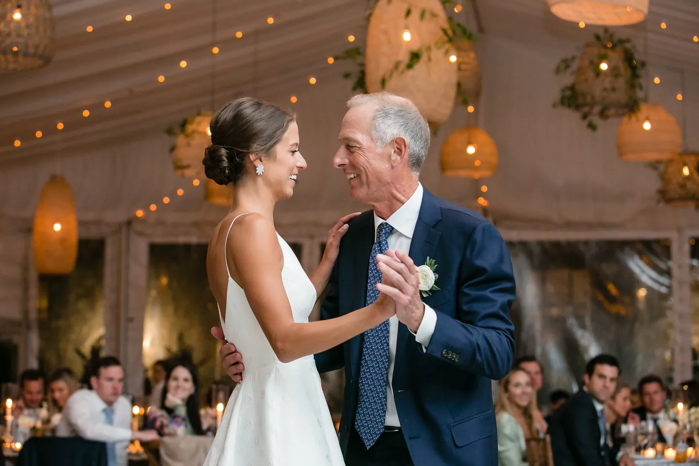 The bride dances with an older man in a navy suit beneath string lights and lanterns