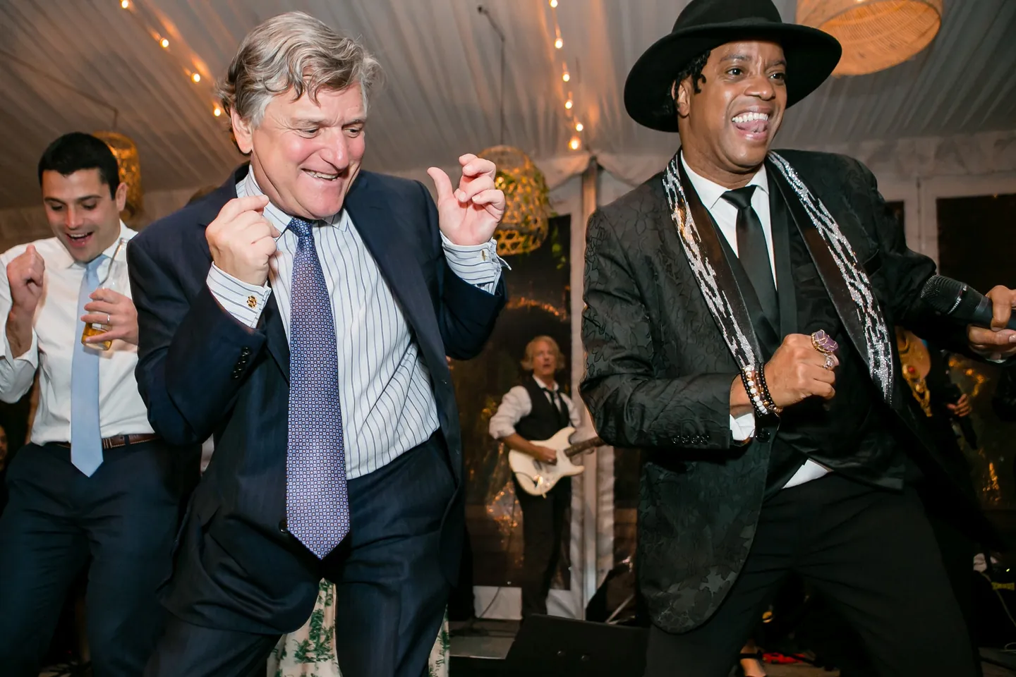 A guest in a tie dances beside a musician in a black hat as the band plays
