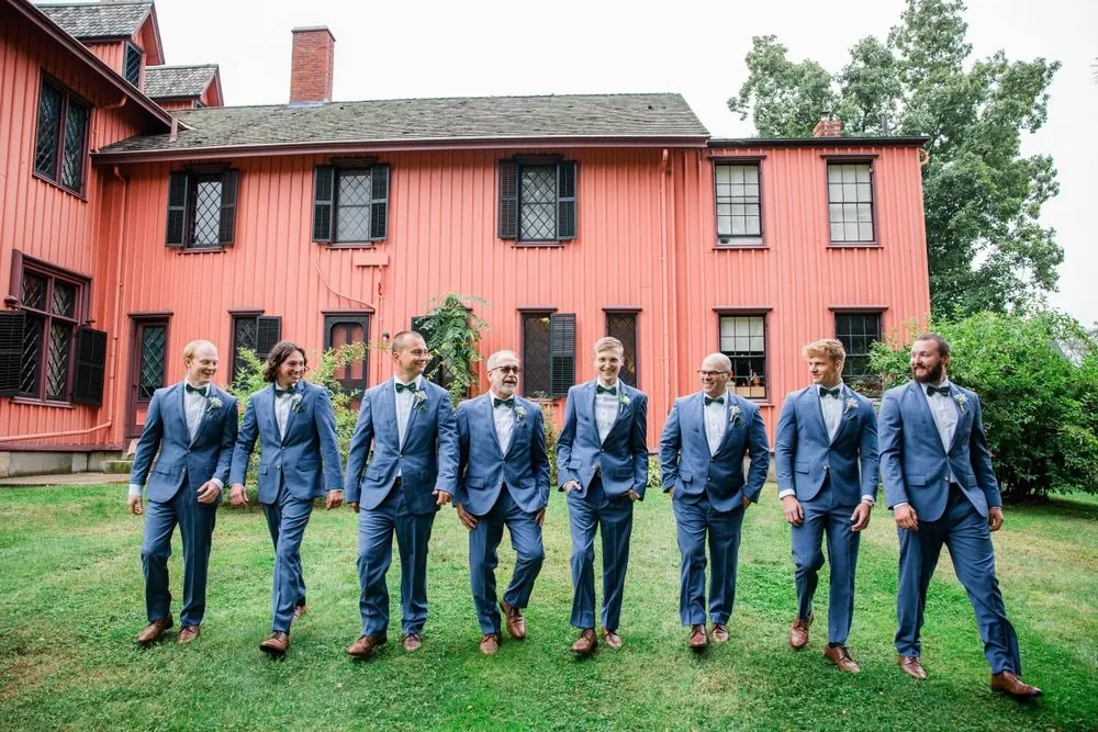 Groomsmen in blue suits walk across the lawn in front of the pink house
