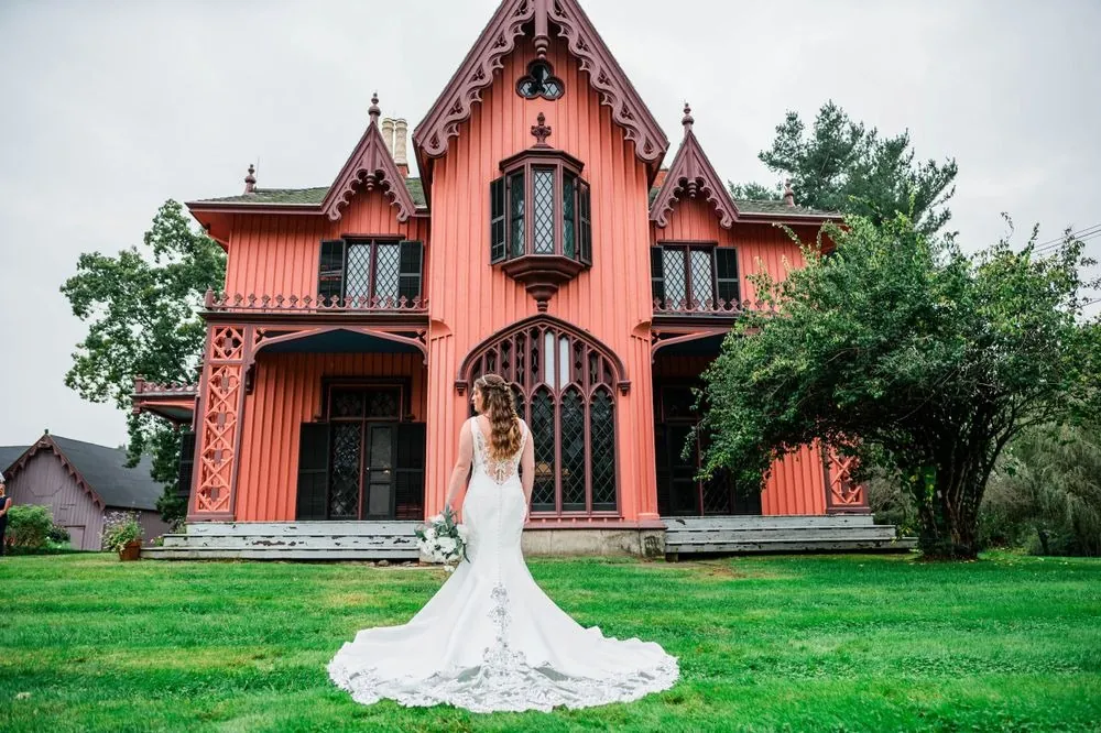 The bride stands on the lawn with her long train spread before the pink cottage