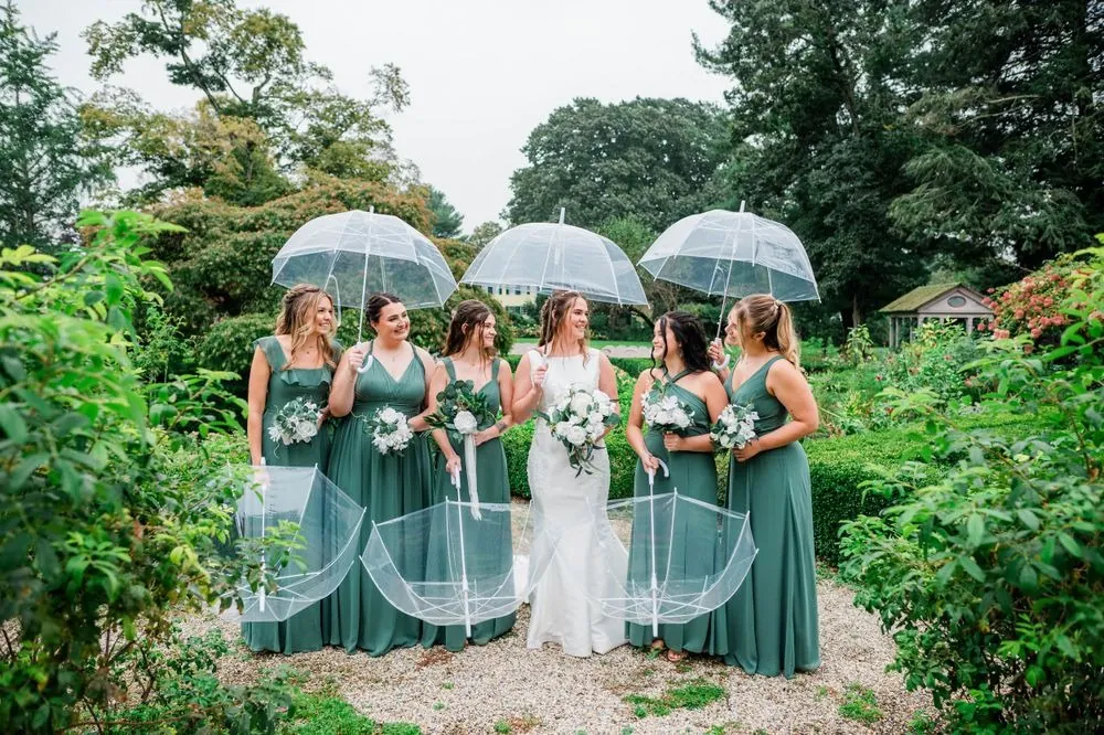 Bridesmaids in sage gowns hold clear umbrellas around the bride in the garden