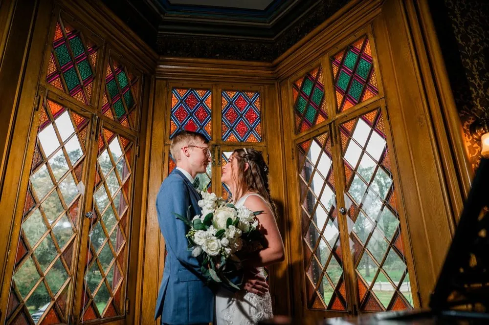 Hunter and Nick stand close inside by colorful diamond-paned stained glass windows