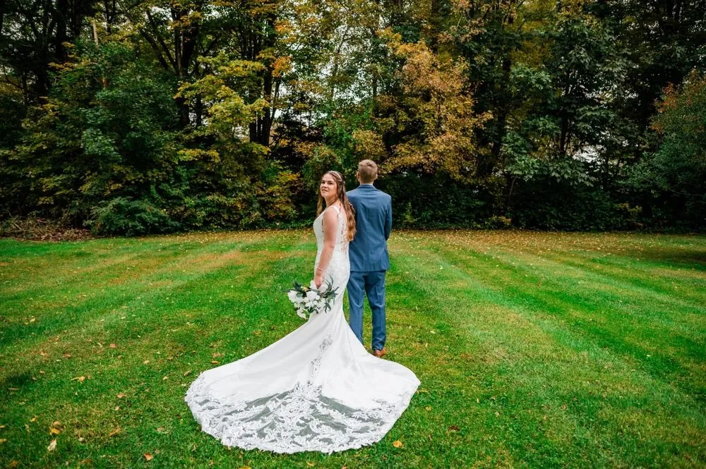 The couple stands on a wide green lawn with her lace train fanned behind her
