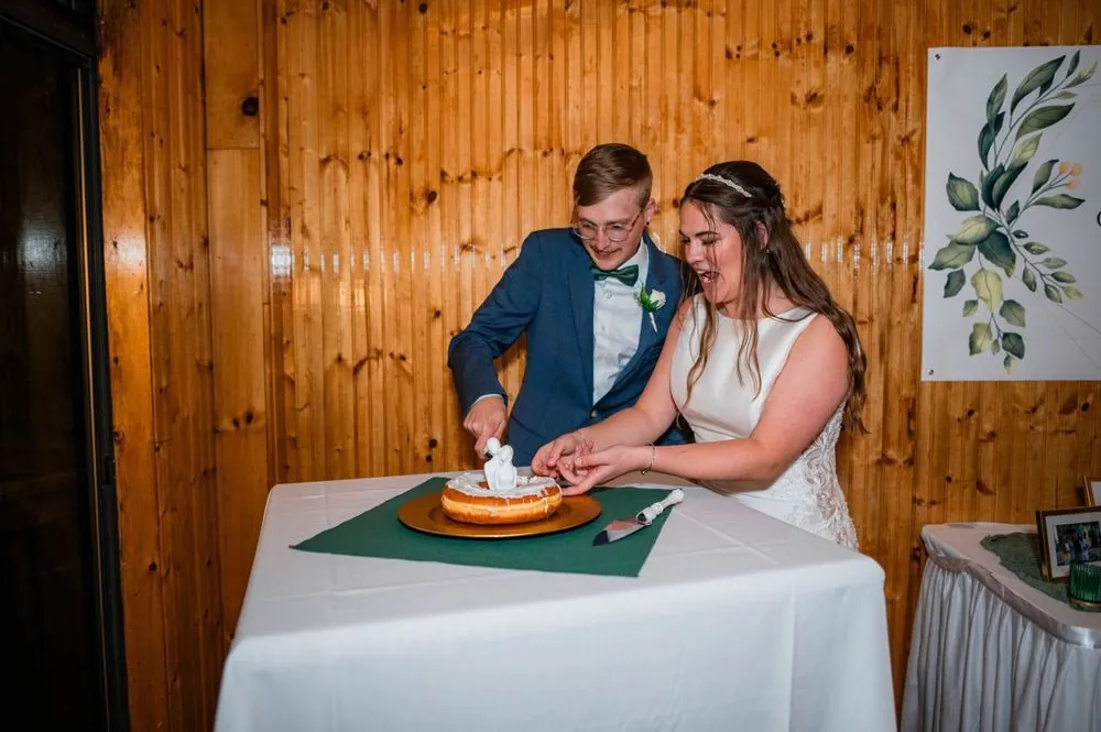 The couple cuts a small round cake at a table against knotty pine walls