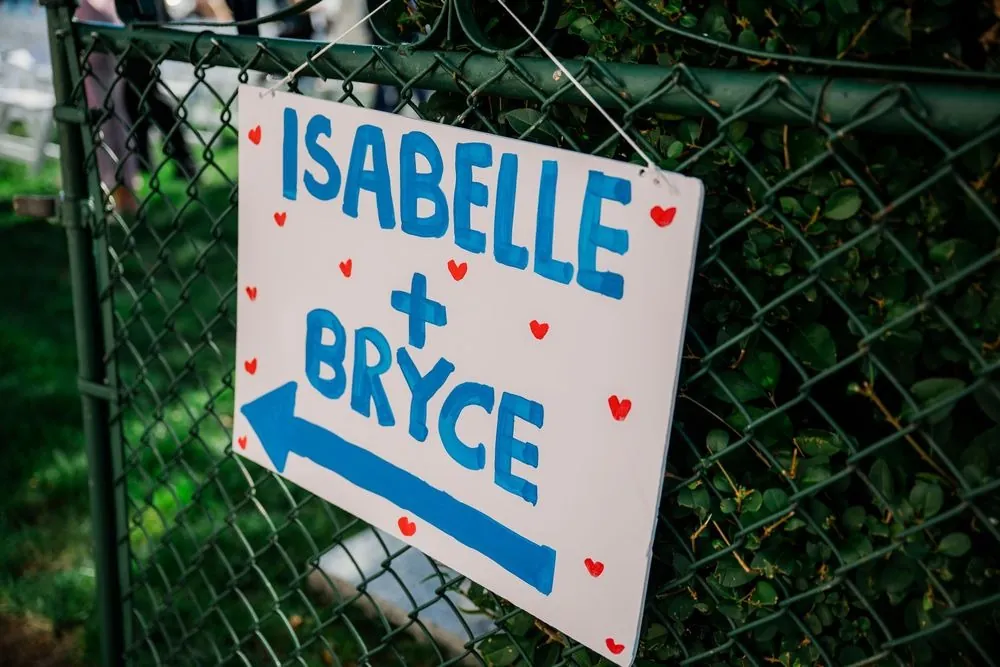 Hand-painted Isabelle and Bryce sign with an arrow, hung on a chain link fence