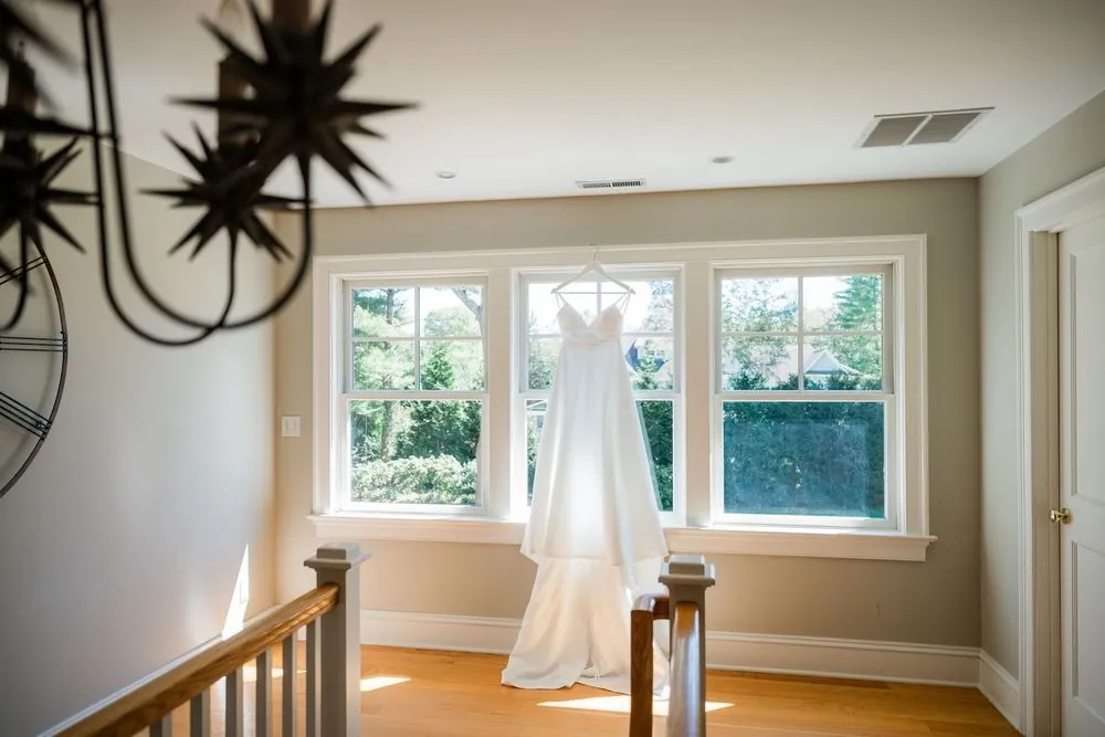Wedding gown hangs by a bright stair landing under a black iron chandelier