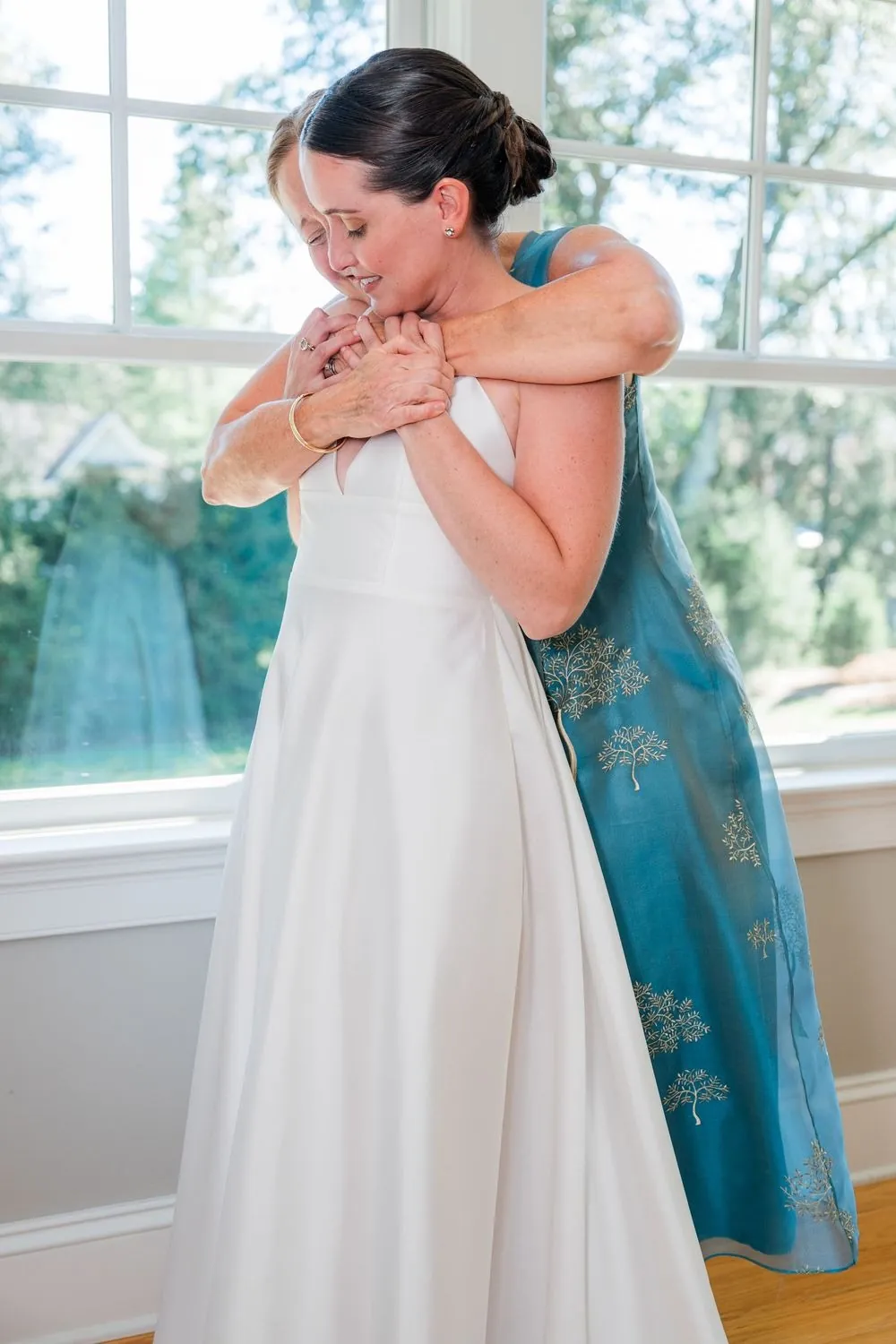 Bride in her gown wraps a woman in a teal dress in a tight hug by the window