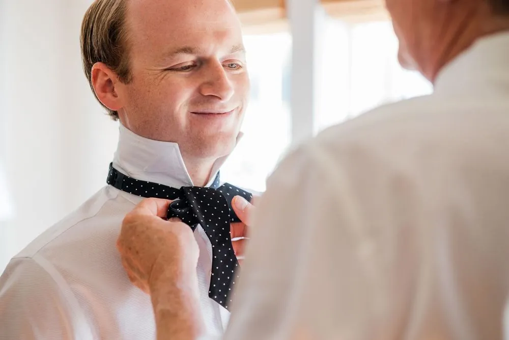 Groom smiles as a hand ties his polka dot bow tie