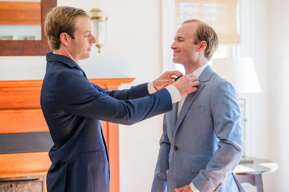 Groomsman in navy straightens the groom's bow tie beside a brick fireplace