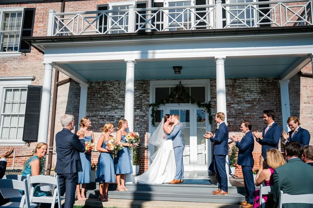 Isabelle and Bryce kiss on the columned porch as the wedding party applauds