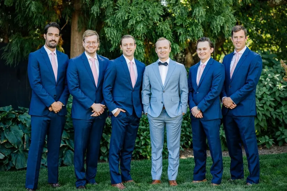 Groomsmen in navy suits stand around the groom in pale blue against green trees