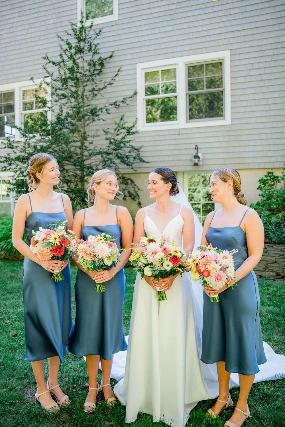 Bridesmaids in slate blue satin hold bright bouquets beside the bride on the lawn