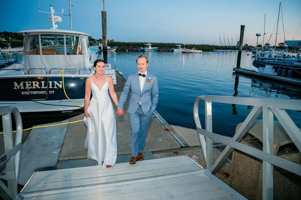 Isabelle and Bryce walk hand in hand down the dock past a moored boat in late light