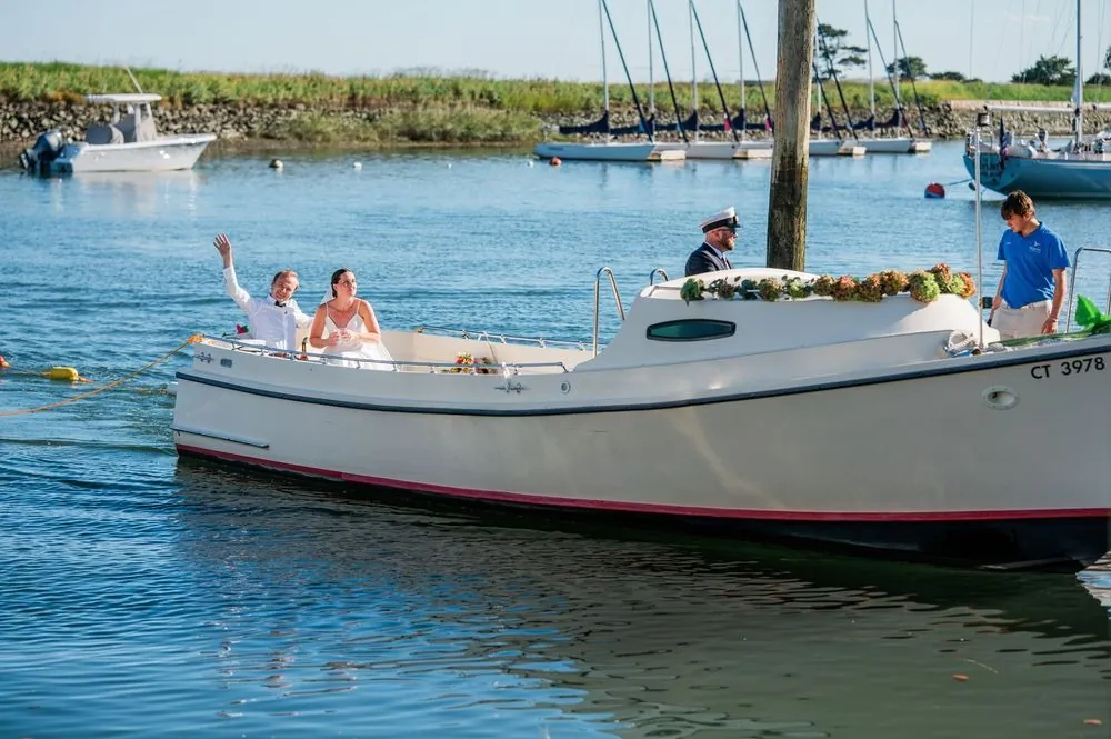 Isabelle and Bryce wave from a white launch cruising past sailboats in the harbor