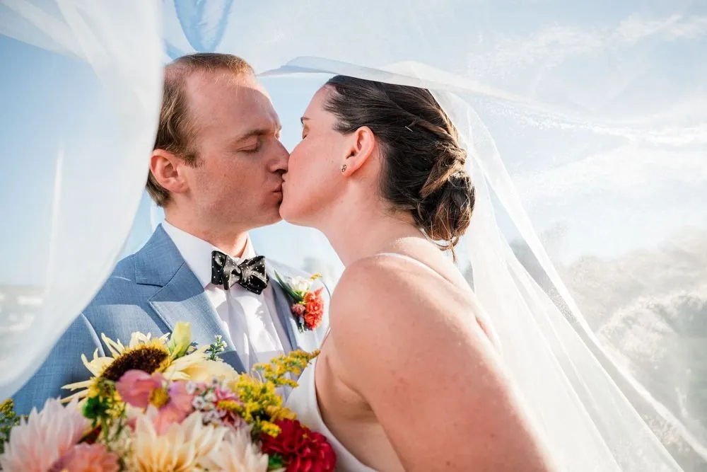 Isabelle and Bryce kiss under her veil with her dahlia and sunflower bouquet in front