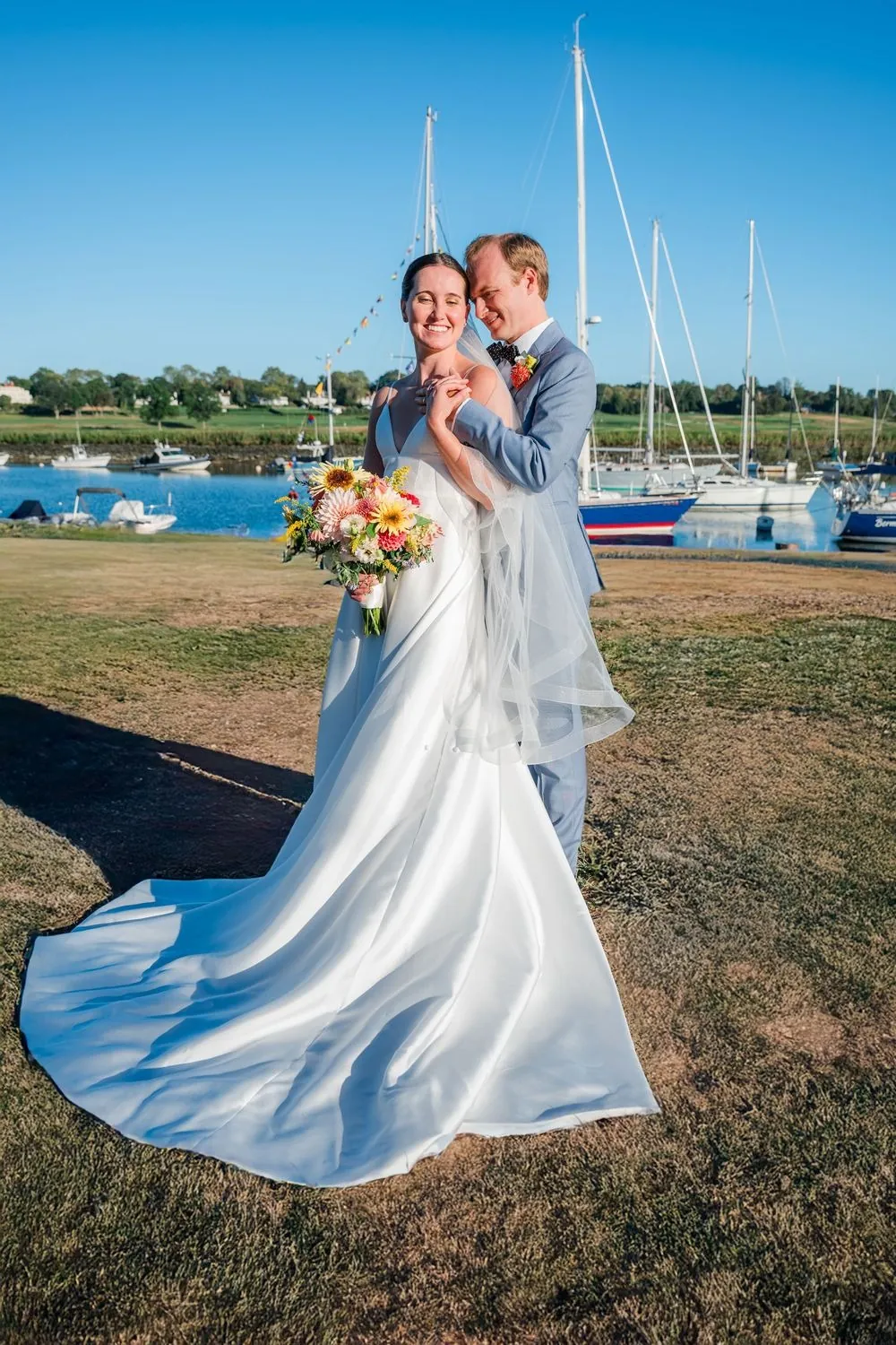 Bryce holds Isabelle close on the harbor lawn, her long train spread on the grass