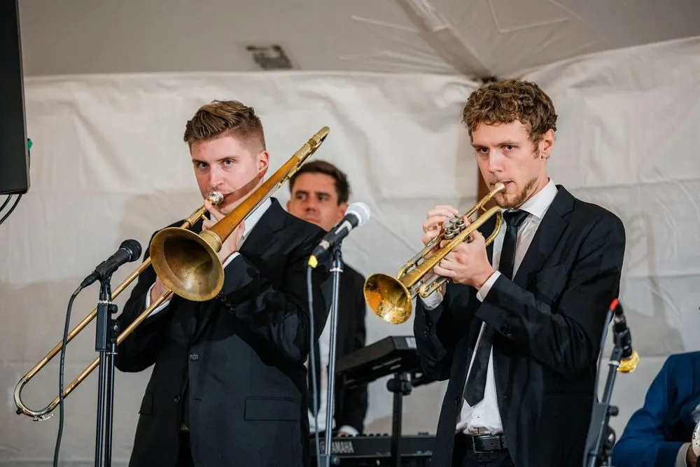 Trombone and trumpet players in dark suits perform under the tent