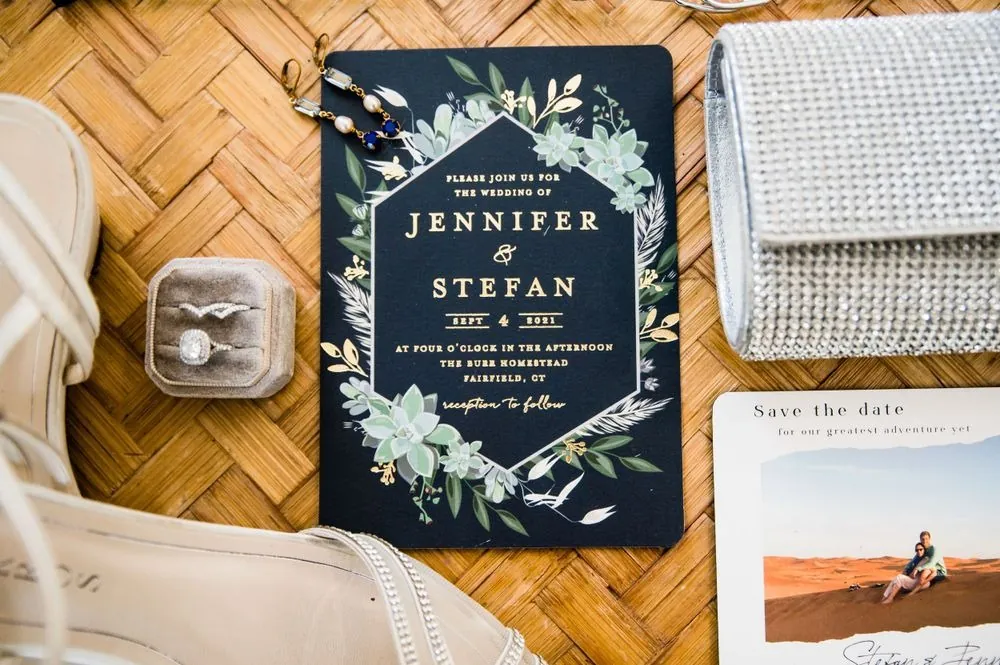 A navy invitation with a gold border and greenery, a ring box and clutch beside it
