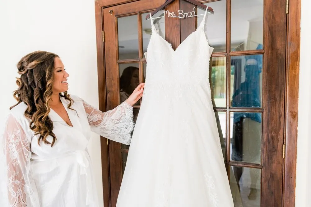 The bride in a lace robe reaches for her gown hanging in a wooden doorway
