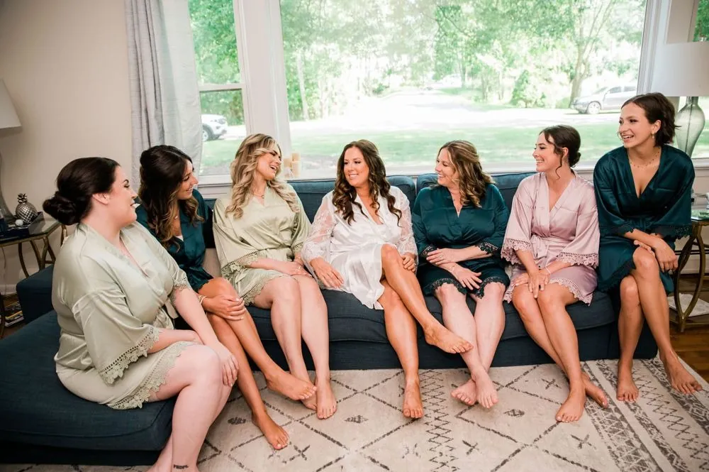 Bridesmaids in satin robes sit together on a sofa in a bright living room