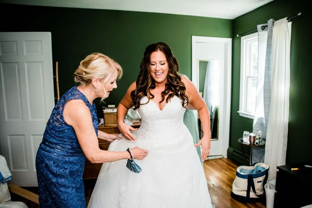 A woman in navy fastens the back of the bride's gown in a dark green room