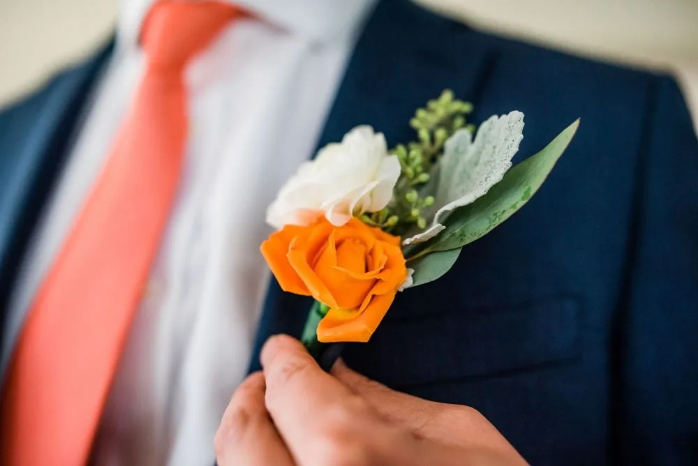 An orange rose boutonniere pinned to a navy lapel beside a coral tie