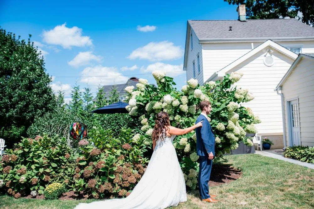 The couple meets beside a tall hedge of white hydrangeas under a blue sky