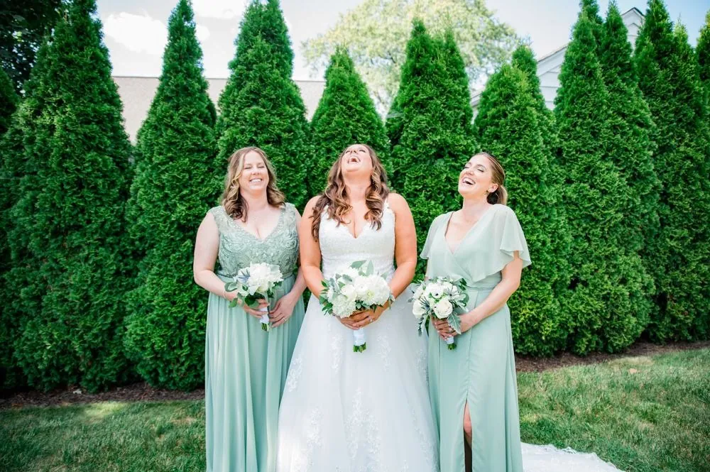 The bride laughs with bridesmaids in sage chiffon gowns against green hedges