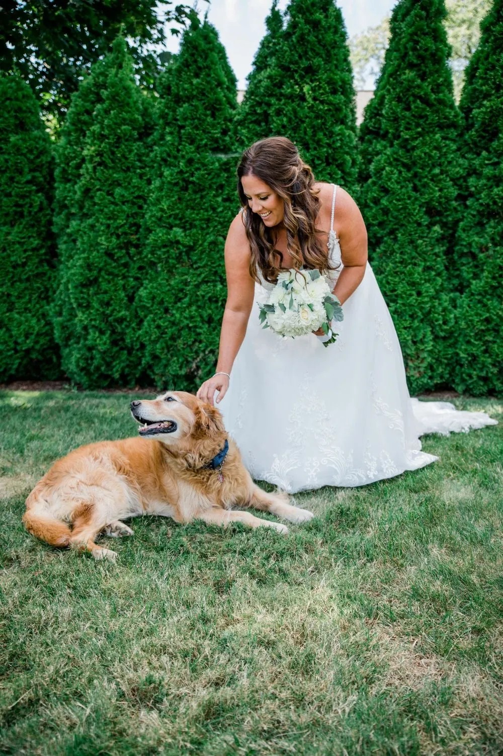 The bride bends to pet the golden retriever on the lawn