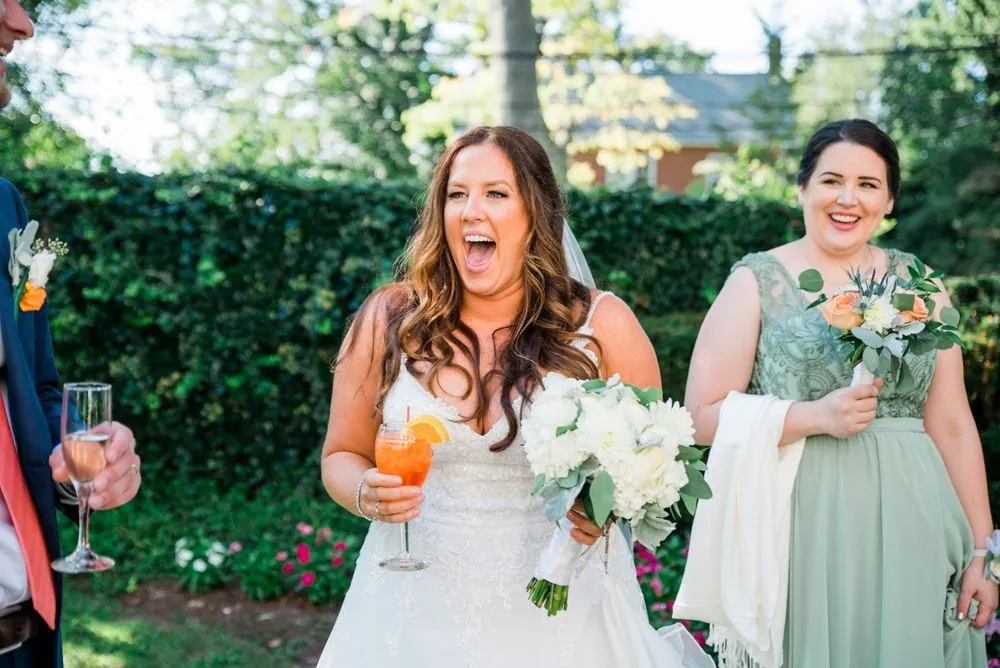 The bride laughs with a drink in hand during cocktail hour on the lawn