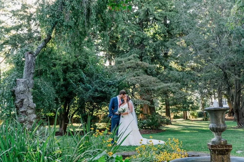 Jennifer and Stefan kiss in the garden under tall old trees beside a stone urn
