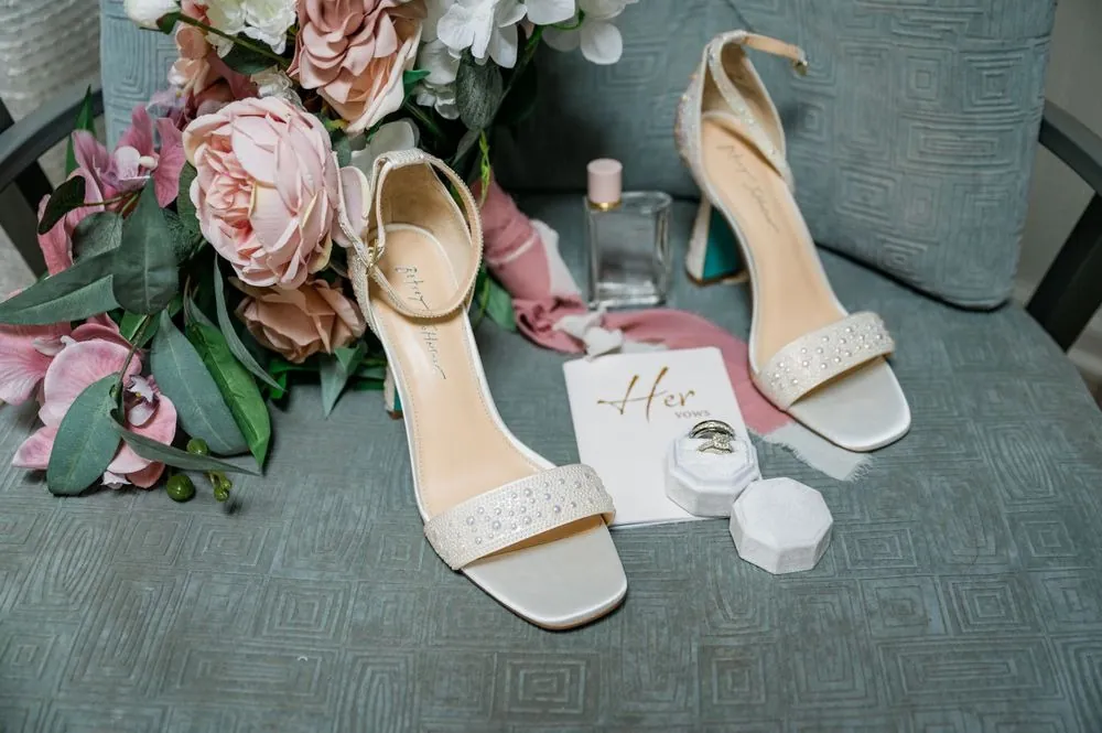Strappy nude heels on a gray chair beside blush roses and a vow booklet