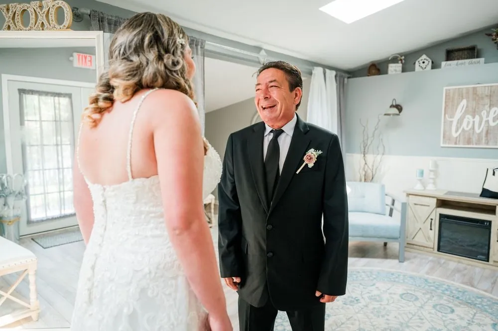 An older man in a dark suit laughs with delight seeing the bride in her gown