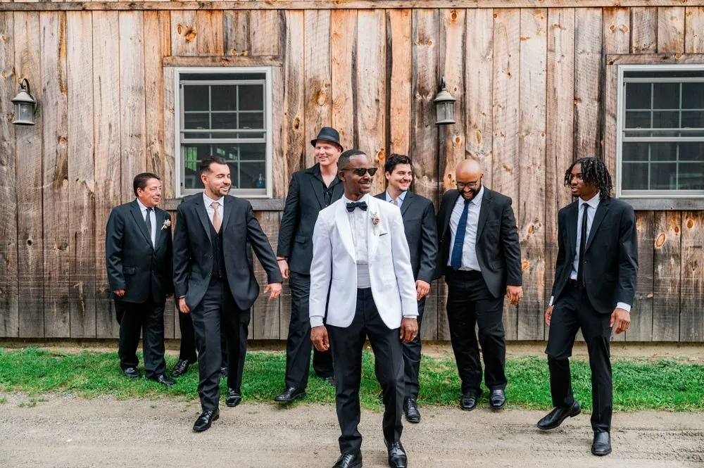 The groom in a white dinner jacket walks with groomsmen in black past a weathered barn
