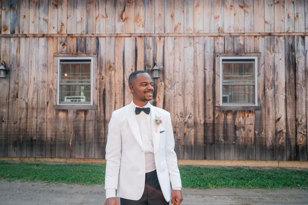 The groom stands against weathered barn siding in a white jacket and bow tie