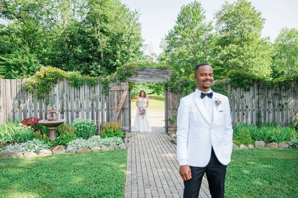 The groom waits on a brick garden path lined with greenery before the first look