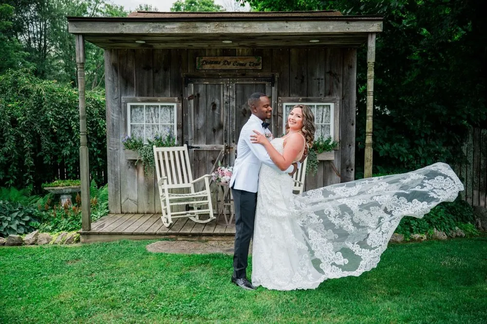 Tifany's lace train flares in the wind as Jhamar holds her by a rustic garden shed