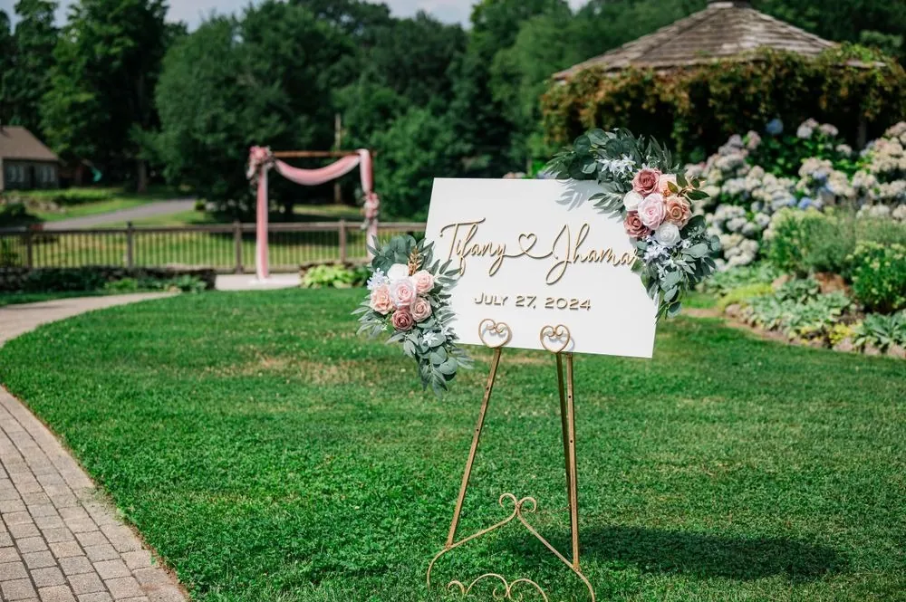 Welcome sign with blush roses on a gold easel, the draped arch across the lawn