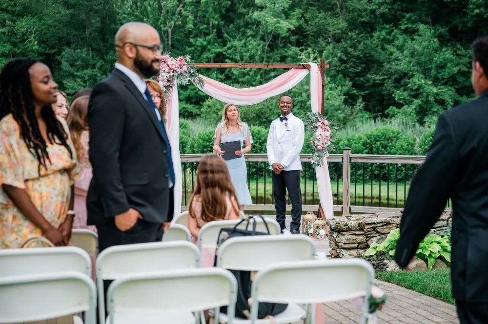 The groom and officiant wait under the blush-draped arch as guests stand