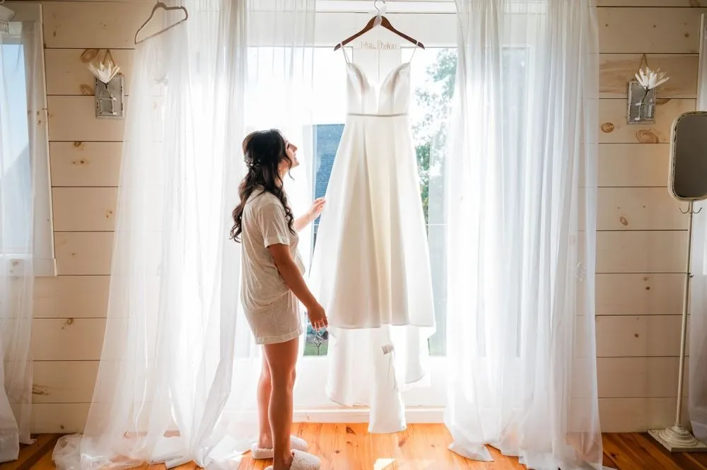 The bride in a white robe looks at her gown hanging in a window of sheer curtains