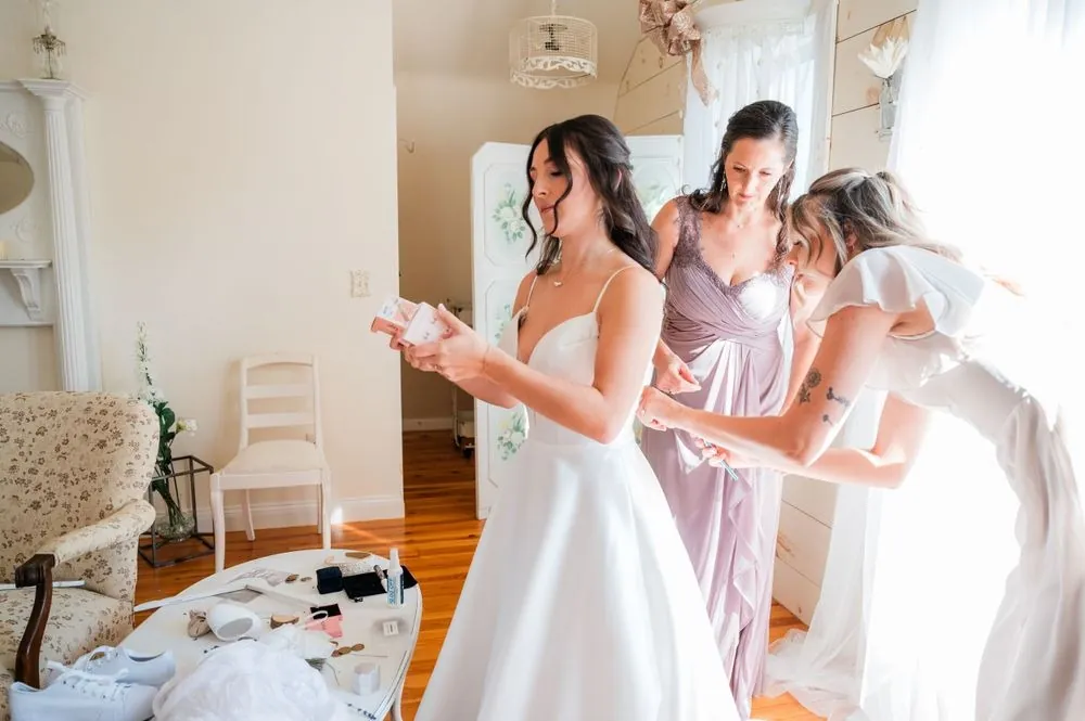 Bridesmaids in blush and mauve fasten the bride's satin gown by the window