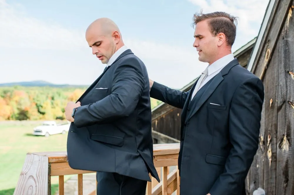 A groomsman helps the groom into his black suit jacket outside a wood building